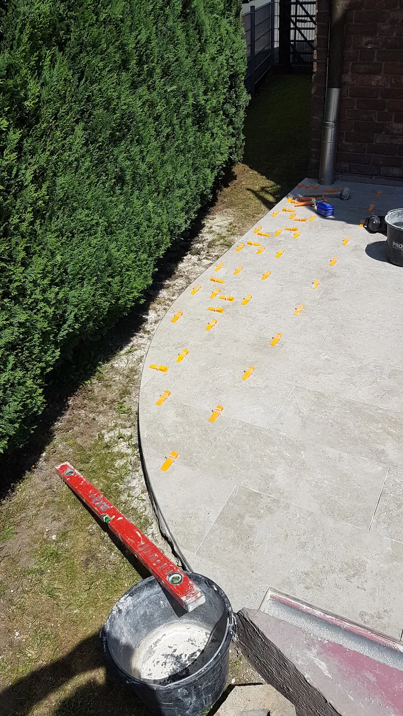 Runde Terrasse mit frisch verlegten hellen Fliesen und Nivellierklammern an einer Hecke