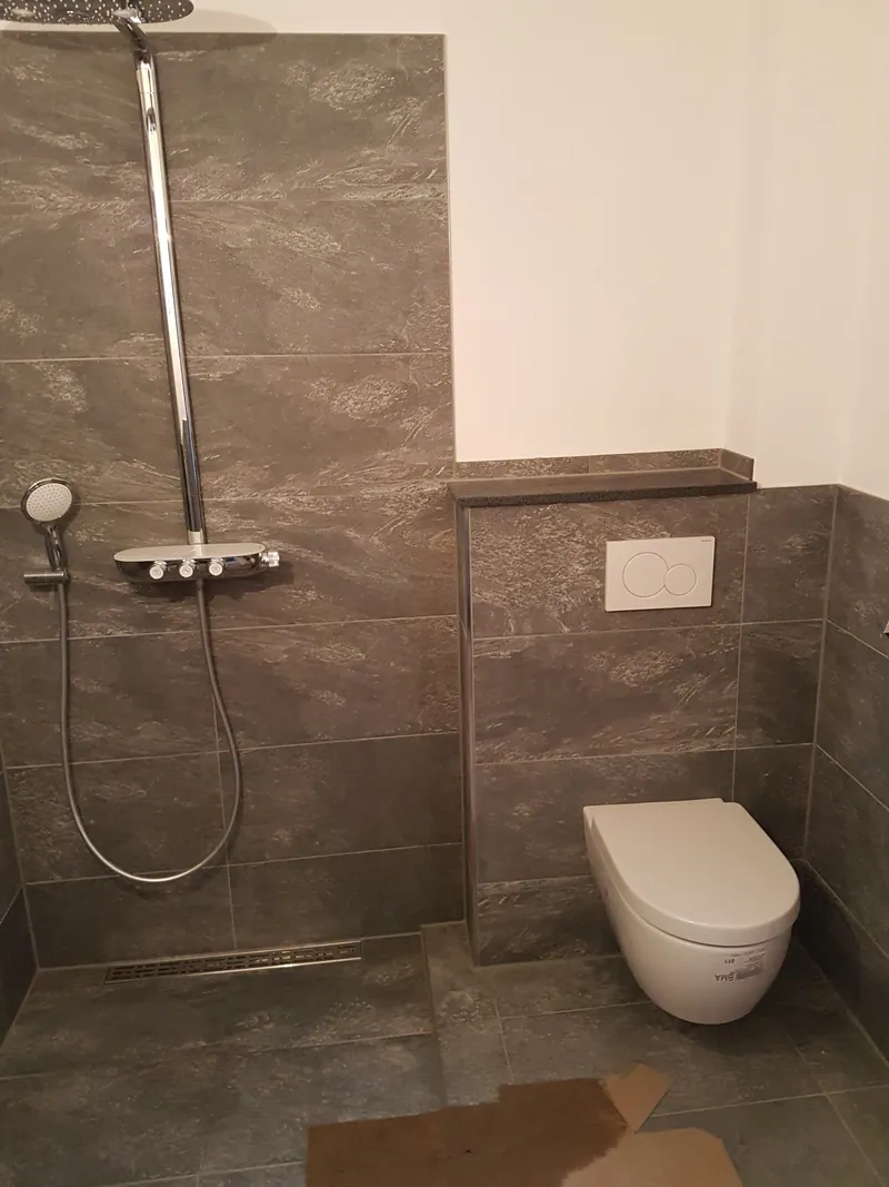 Bad mit großformatigen braungrauen Wandfliesen, bodengleicher Dusche und Vorwand-WC