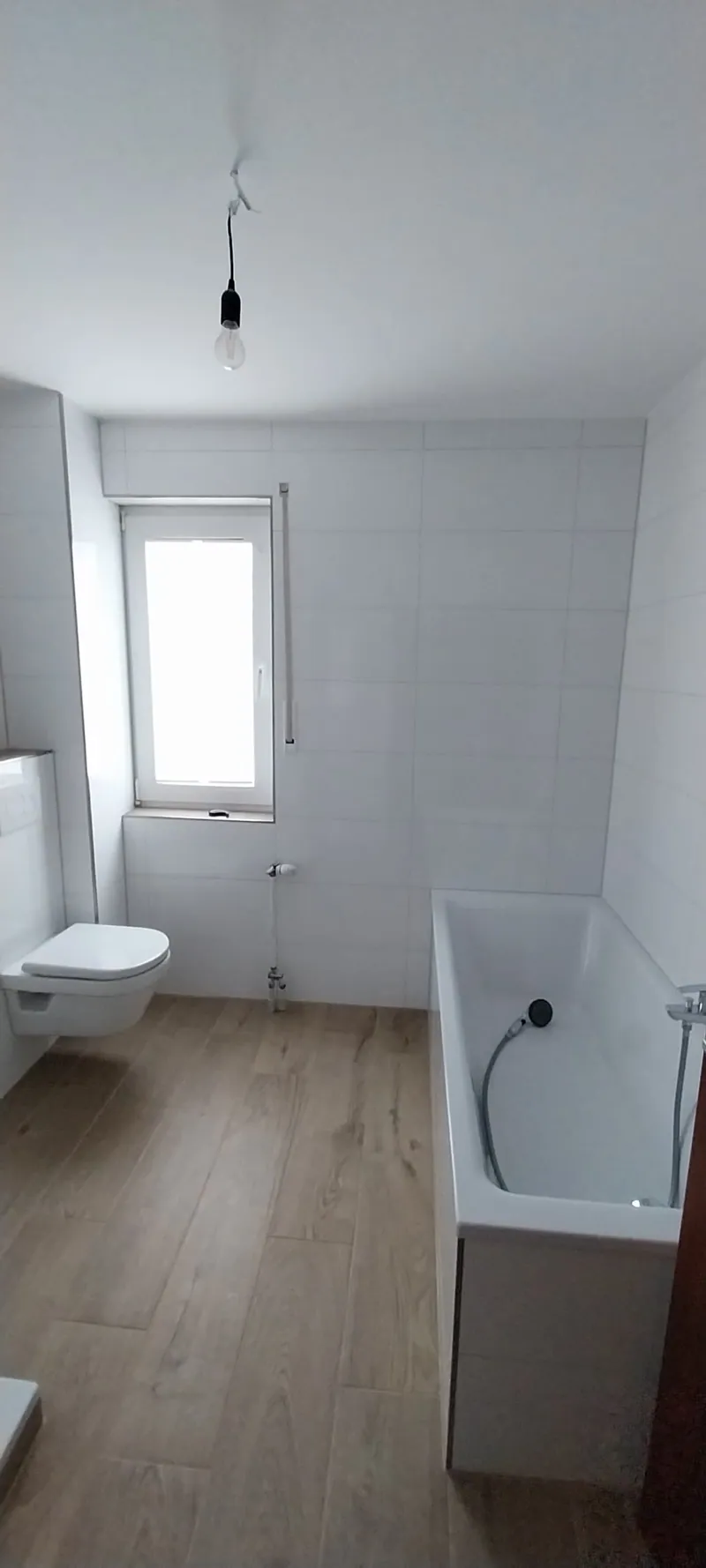 Schmales Bad mit weißen Wandfliesen, Badewanne und Holzoptik-Boden