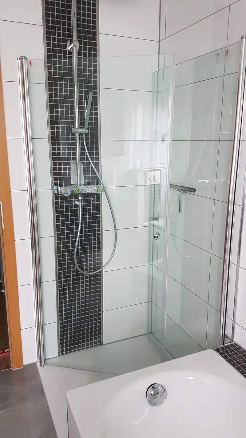 Dusche mit weißen Wandfliesen, schwarzem Mosaikstreifen und Glaswand neben der Badewanne